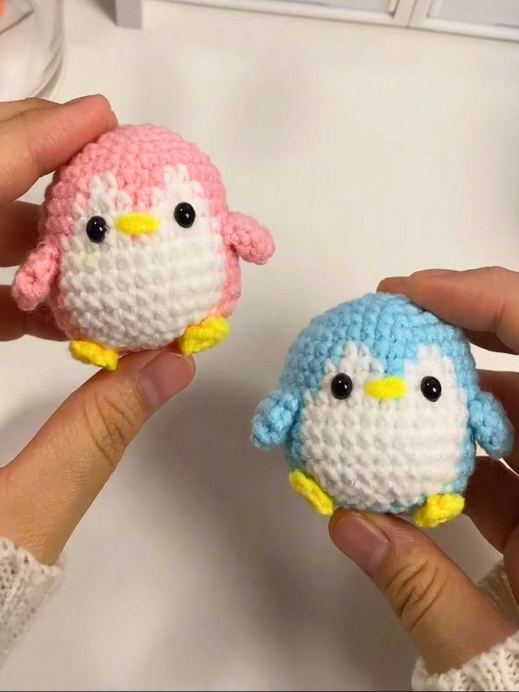 Two small crocheted penguin toys, one pink and one blue, held by hands against a white background.