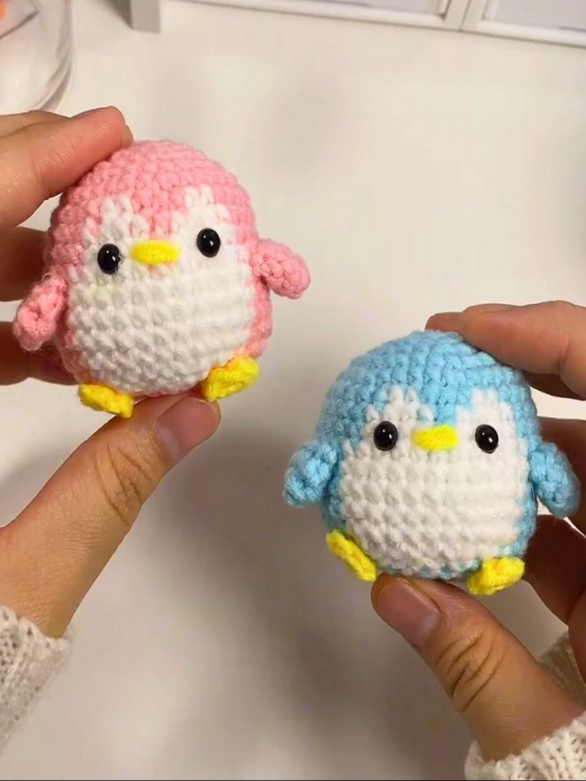 Two small crocheted penguin toys, one pink and one blue, held by hands against a white background.
