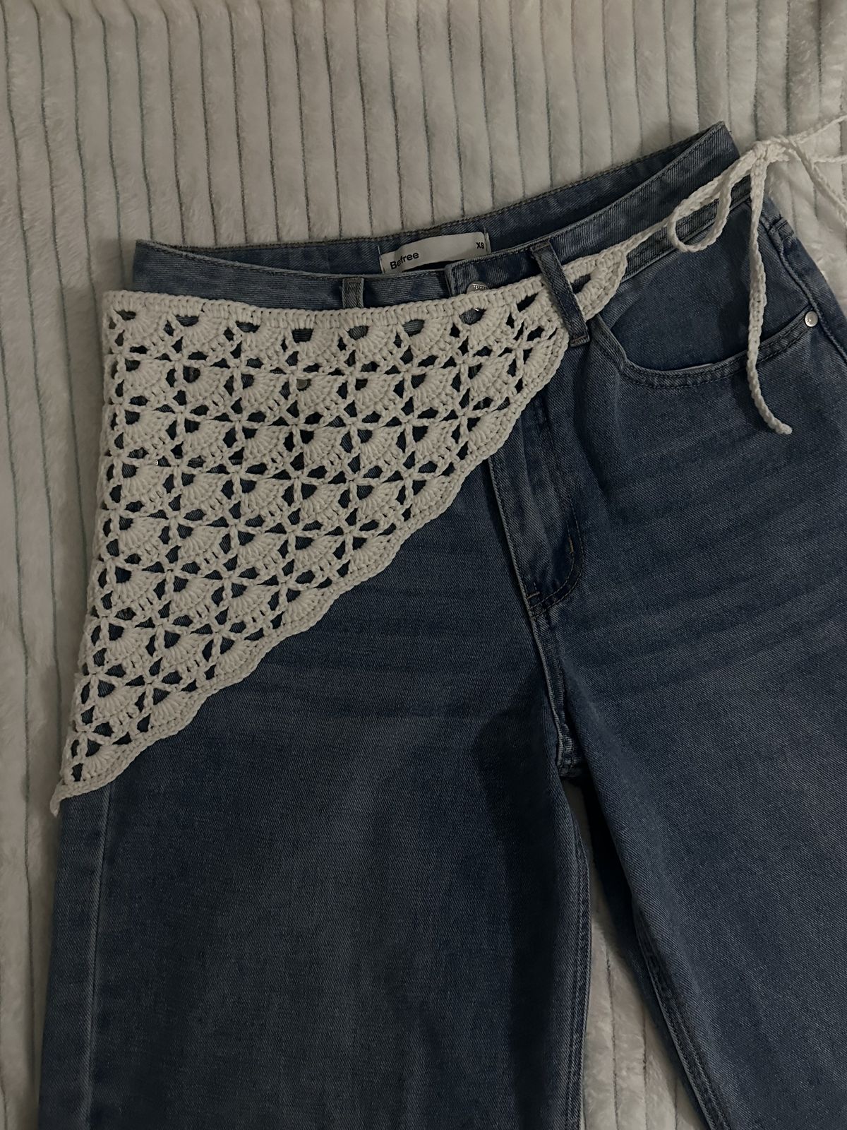 Dark blue jeans with a beige crochet triangle pattern draped over them on a textured surface.