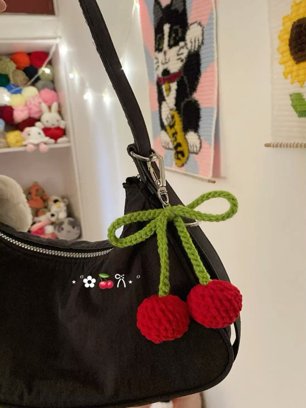 Black handbag with a green and red cherry-themed keychain in a room with stuffed animals and artwork.