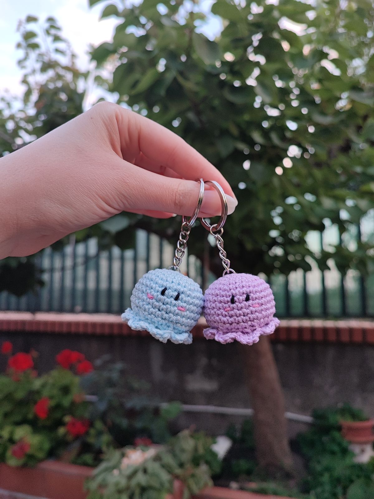 Hand holding two small crocheted keychains shaped like an octopus, one blue and one pink, with a garden background.