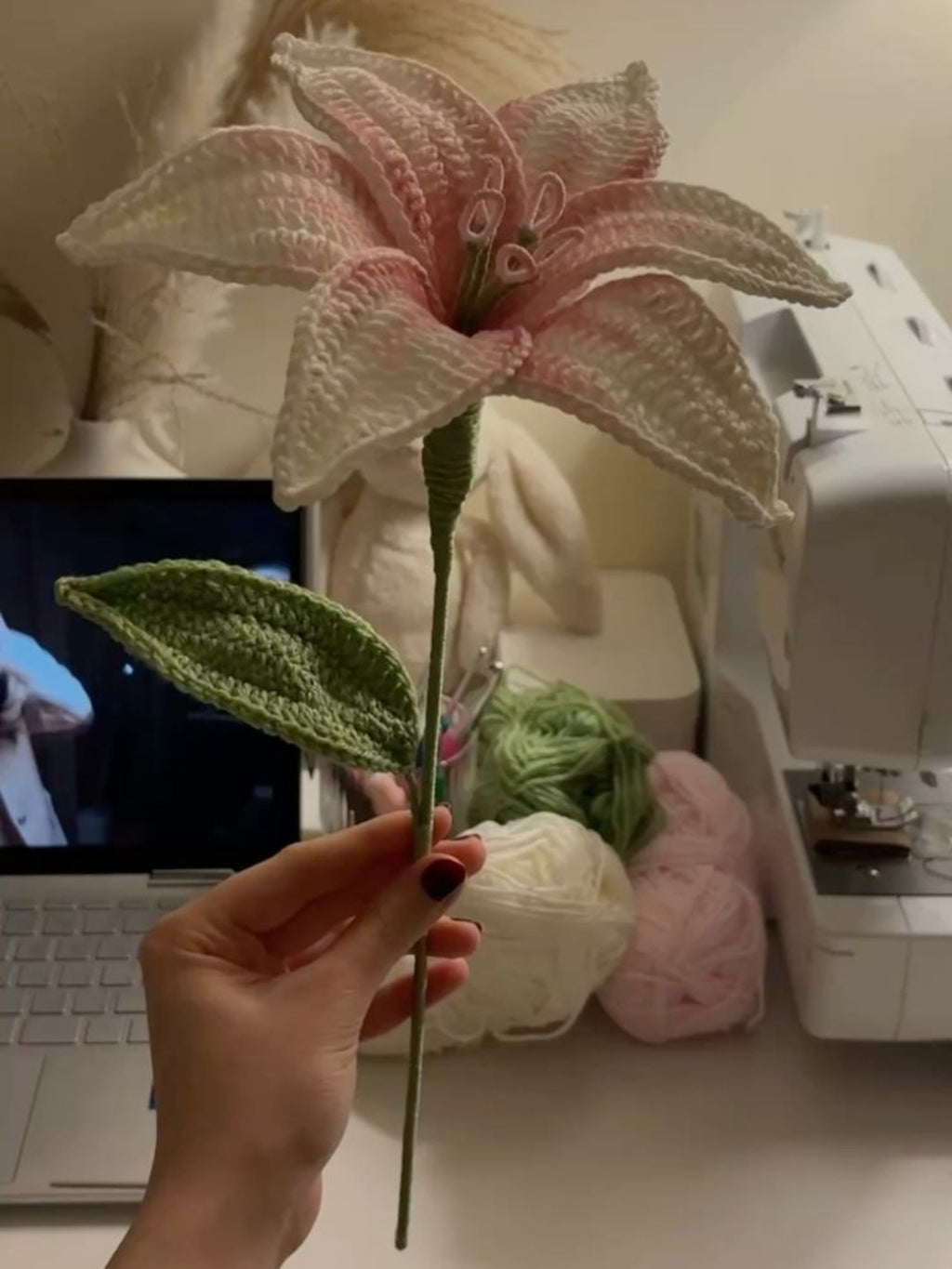 Hand holding a pink artificial flower in front of a sewing machine and computer on a desk.