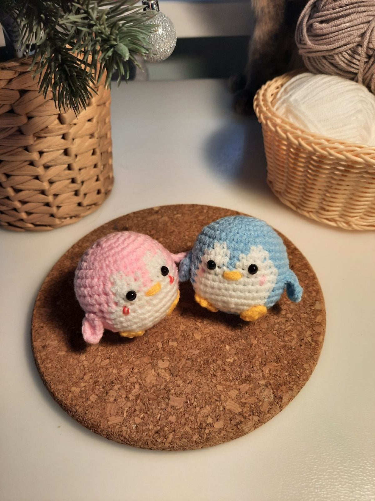 Two small crocheted penguin toys on a cork coaster with wicker baskets in the background.