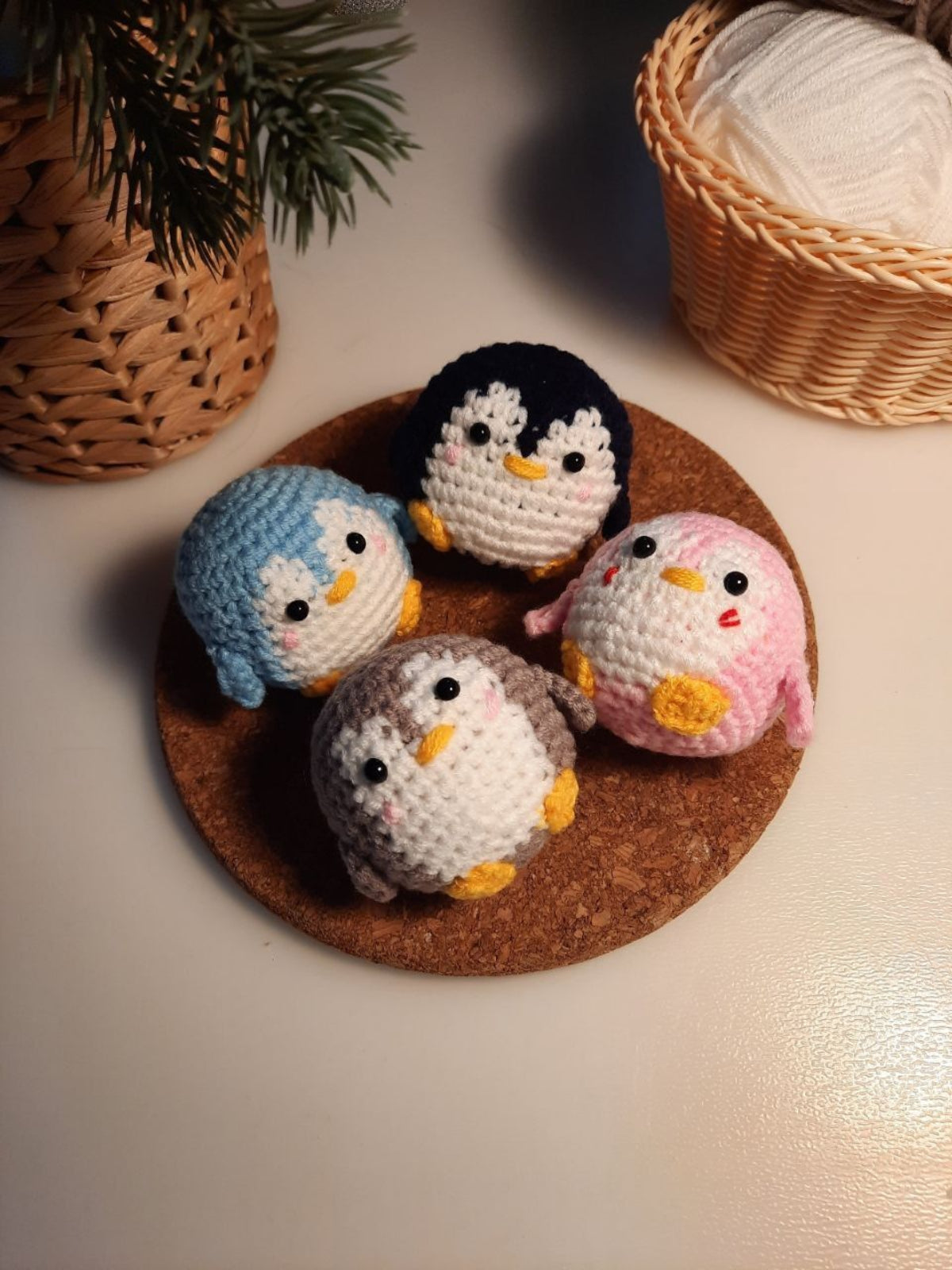 Four crocheted penguin toys on a cork coaster with a basket of yarn in the background.