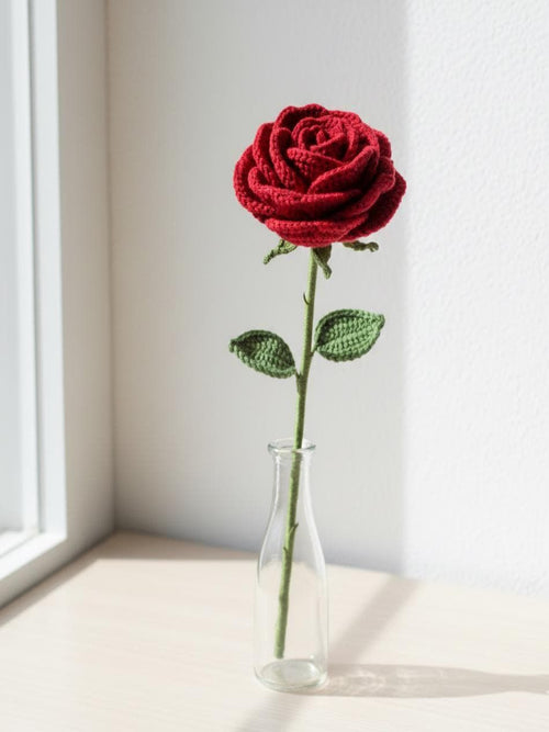 Knitted red rose in a clear vase on a light surface with a window in the background