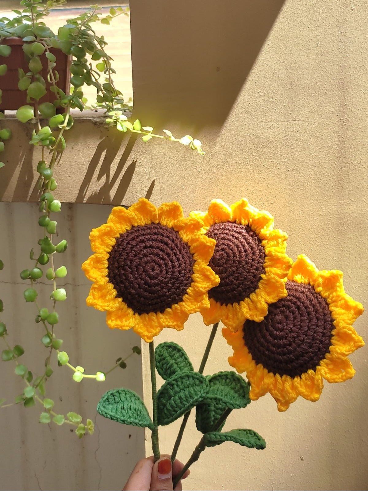 Handmade crochet sunflowers held in front of a window with plants.